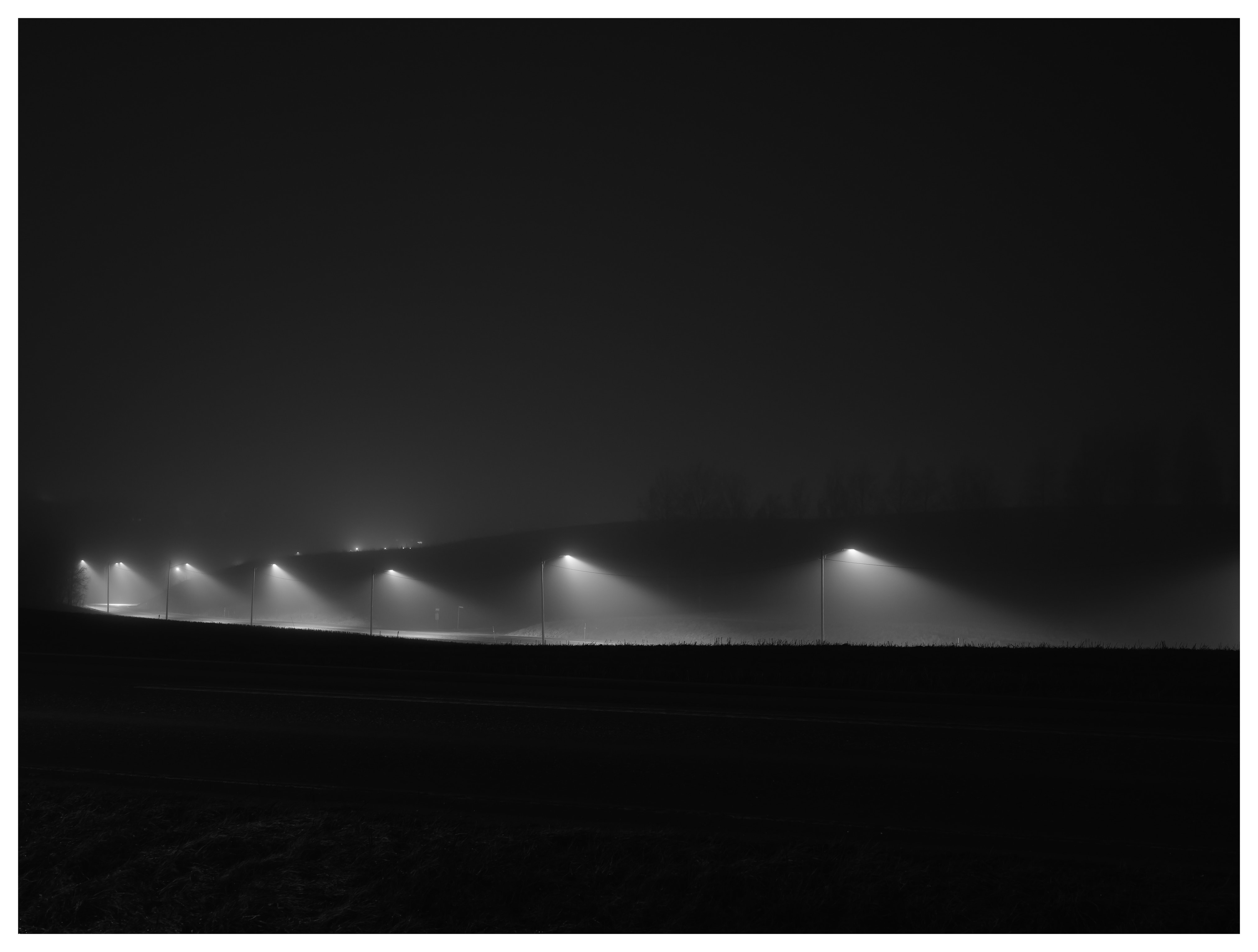 A row of streetlights shining through dense fog at night, casting long beams of light across an empty landscape in black and white.