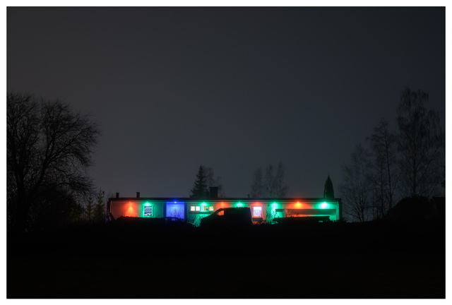 A long, low house illuminated by red, green, and blue lights on a foggy night, with trees and a parked vehicle in silhouette against the dark sky.