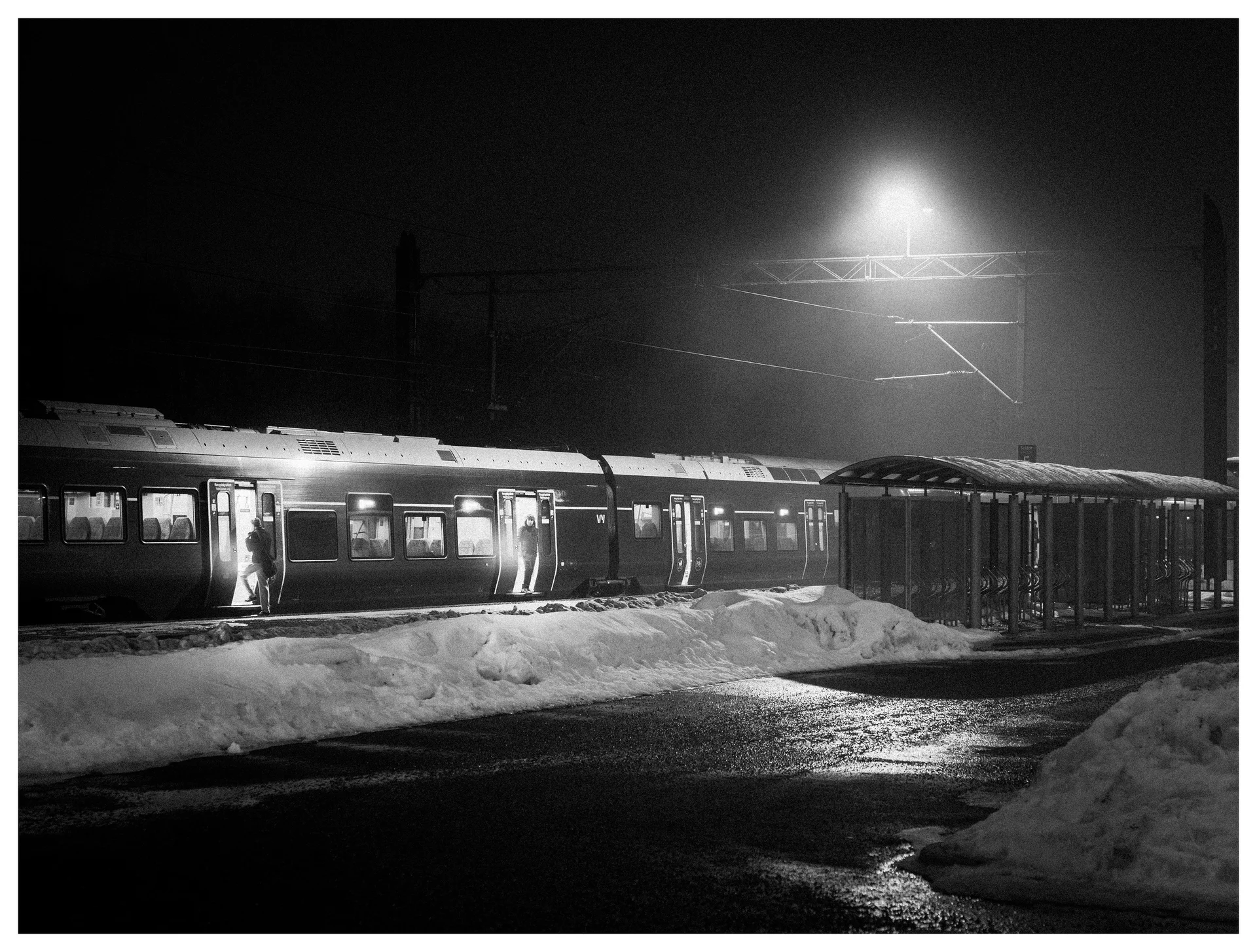 Nighttime black and white photograph of passengers boarding a commuter train at a snowy railway platform with wet pavement reflections and strong overhead light.