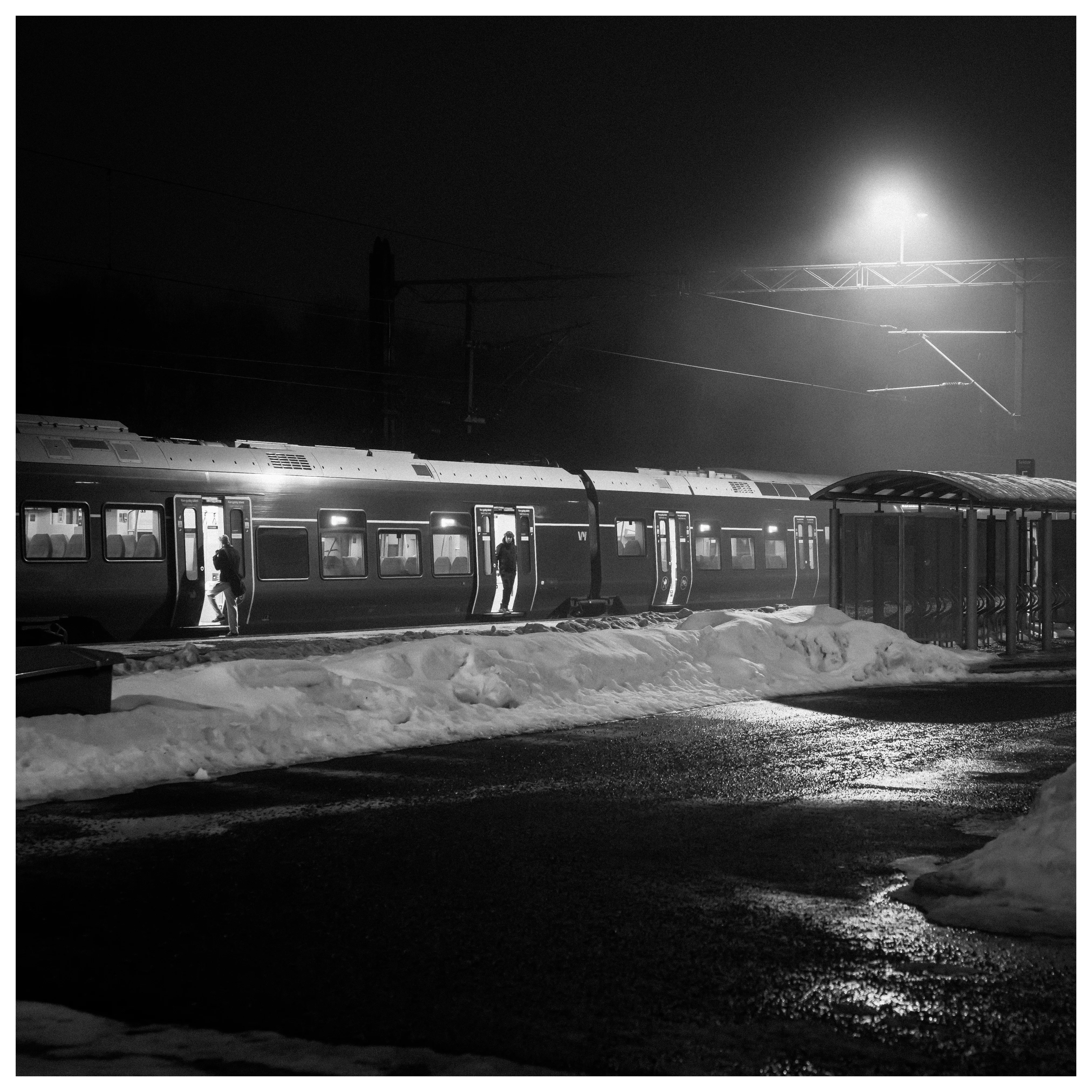 Nighttime black and white photograph of passengers boarding a commuter train at a snowy railway platform with wet pavement reflections and strong overhead light.
