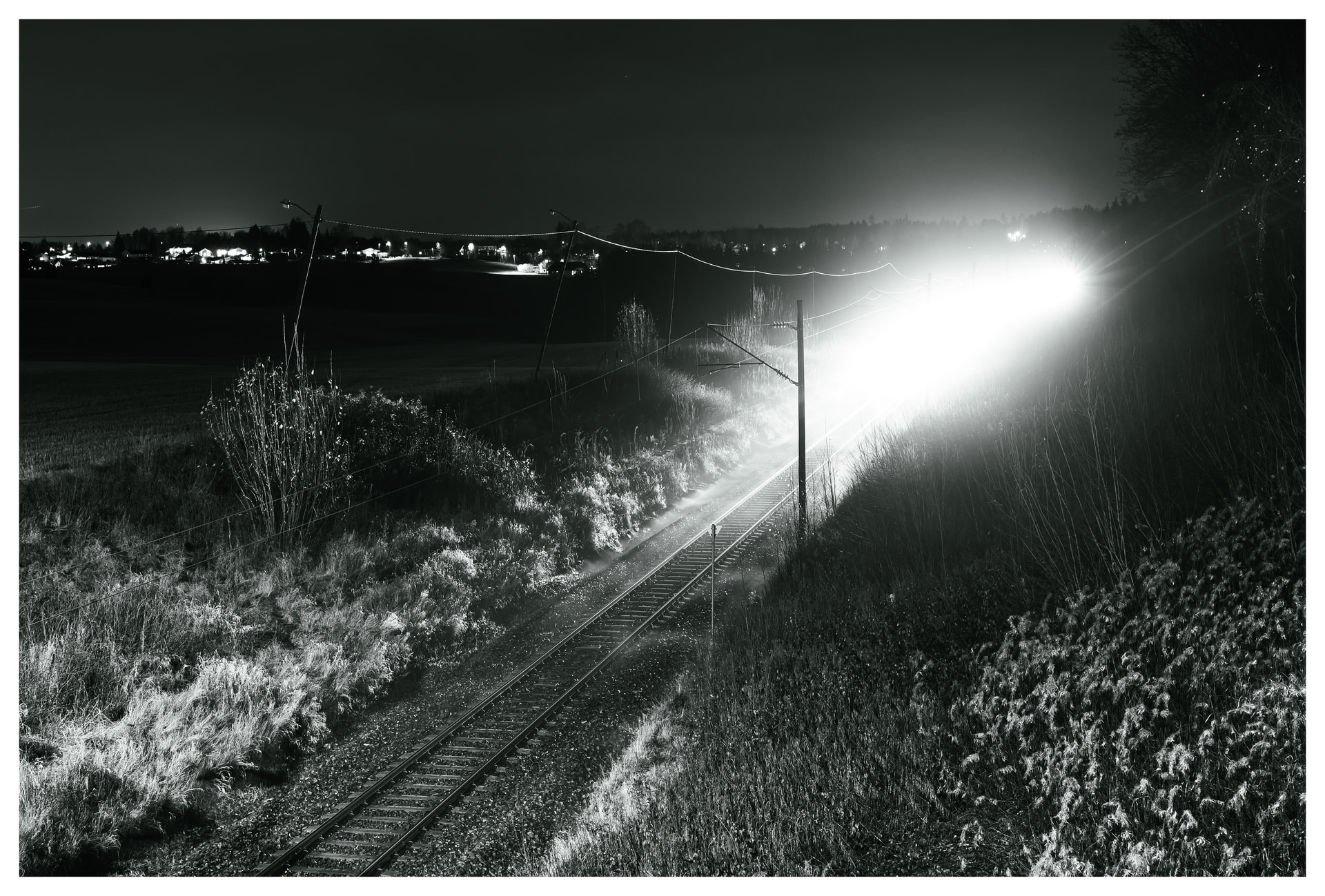 A long-exposure night photo of a bright train moving along rural railway tracks, illuminating grass and landscape as it passes through the dark countryside.