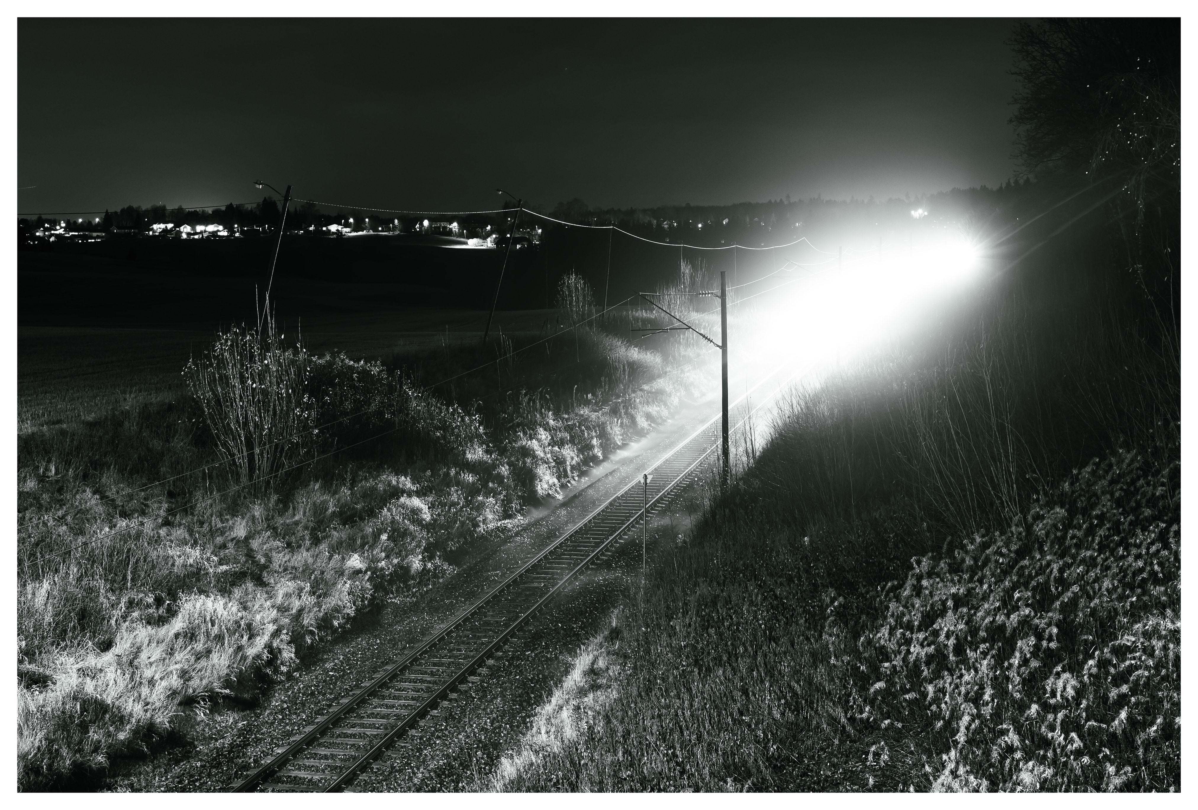 A long-exposure night photo of a bright train moving along rural railway tracks, illuminating grass and landscape as it passes through the dark countryside.
