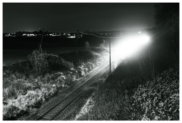A long-exposure night photo of a bright train moving along rural railway tracks, illuminating grass and landscape as it passes through the dark countryside.