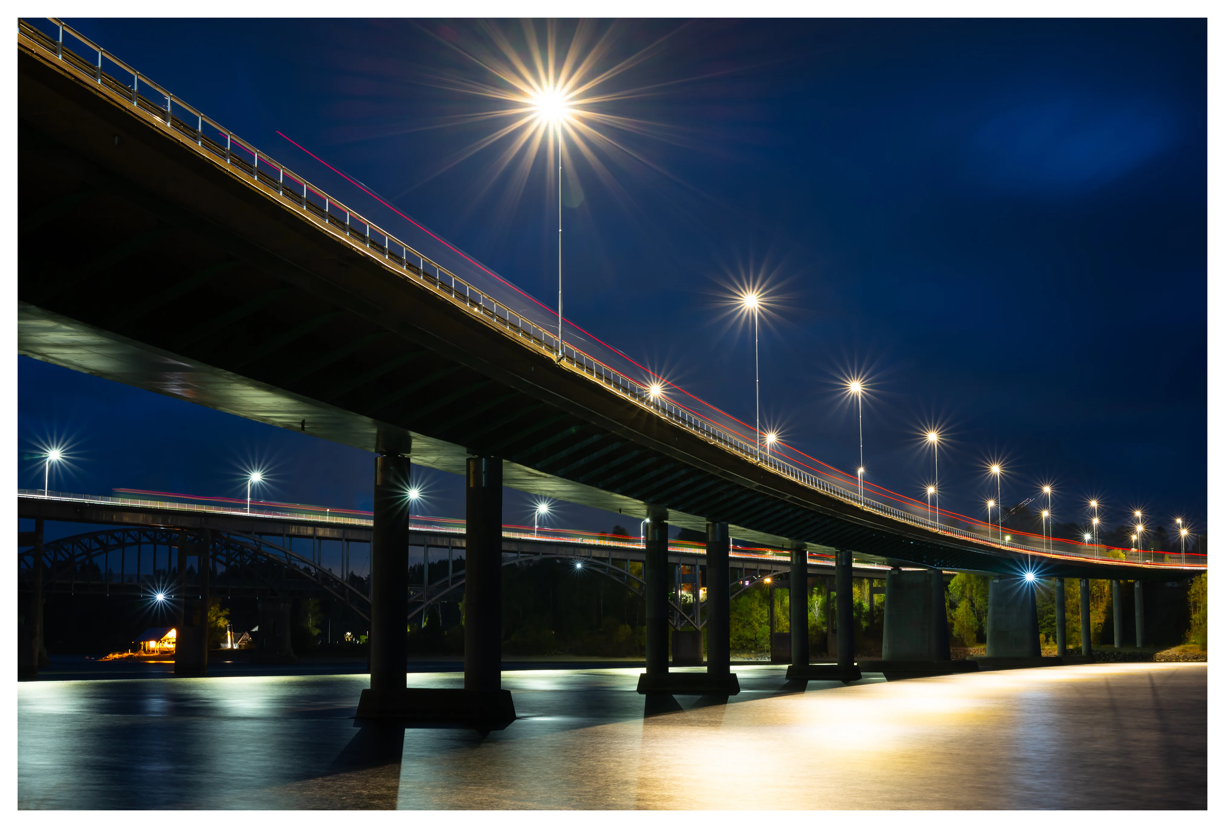 A long bridge at night with bright star-shaped streetlights and red light trails from passing cars, reflected in calm water below.