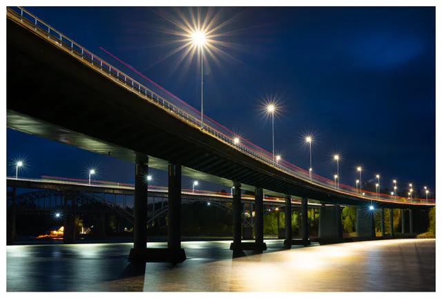 A long bridge at night with bright star-shaped streetlights and red light trails from passing cars, reflected in calm water below.