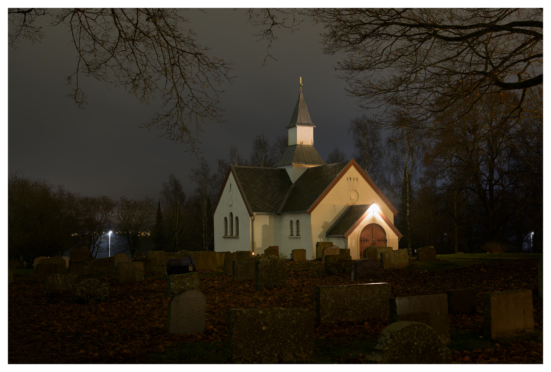 A small white church illuminated by warm lights at night, surrounded by gravestones and bare autumn trees under a cloudy sky.