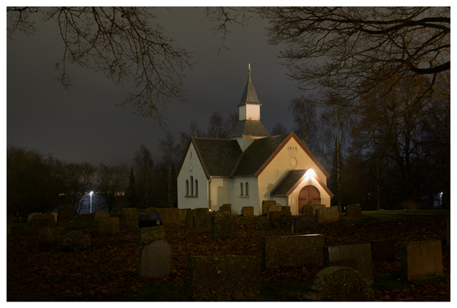 A small white church illuminated by warm lights at night, surrounded by gravestones and bare autumn trees under a cloudy sky.