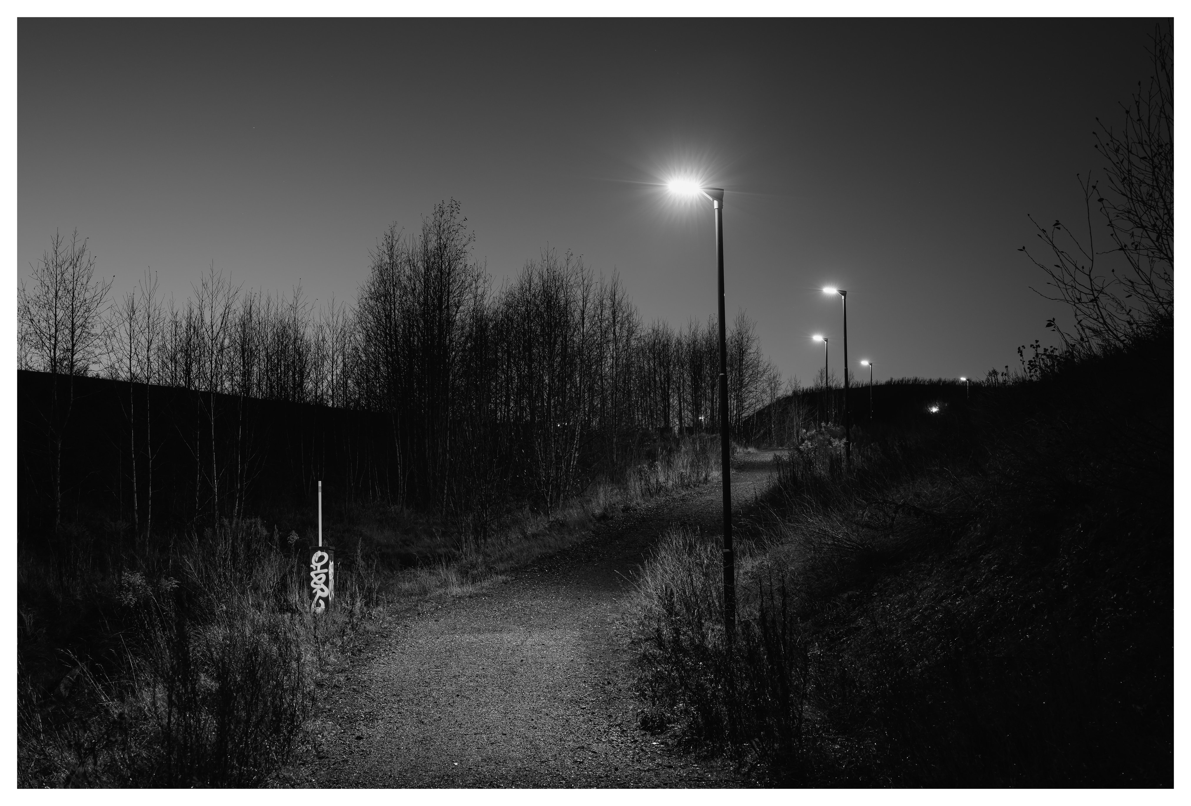 A quiet gravel path at night illuminated by streetlights, with a small graffiti-covered utility post standing among dry grass.