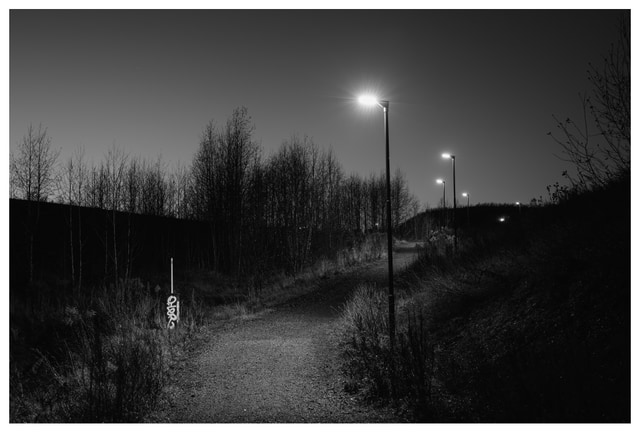 A quiet gravel path at night illuminated by streetlights, with a small graffiti-covered utility post standing among dry grass.