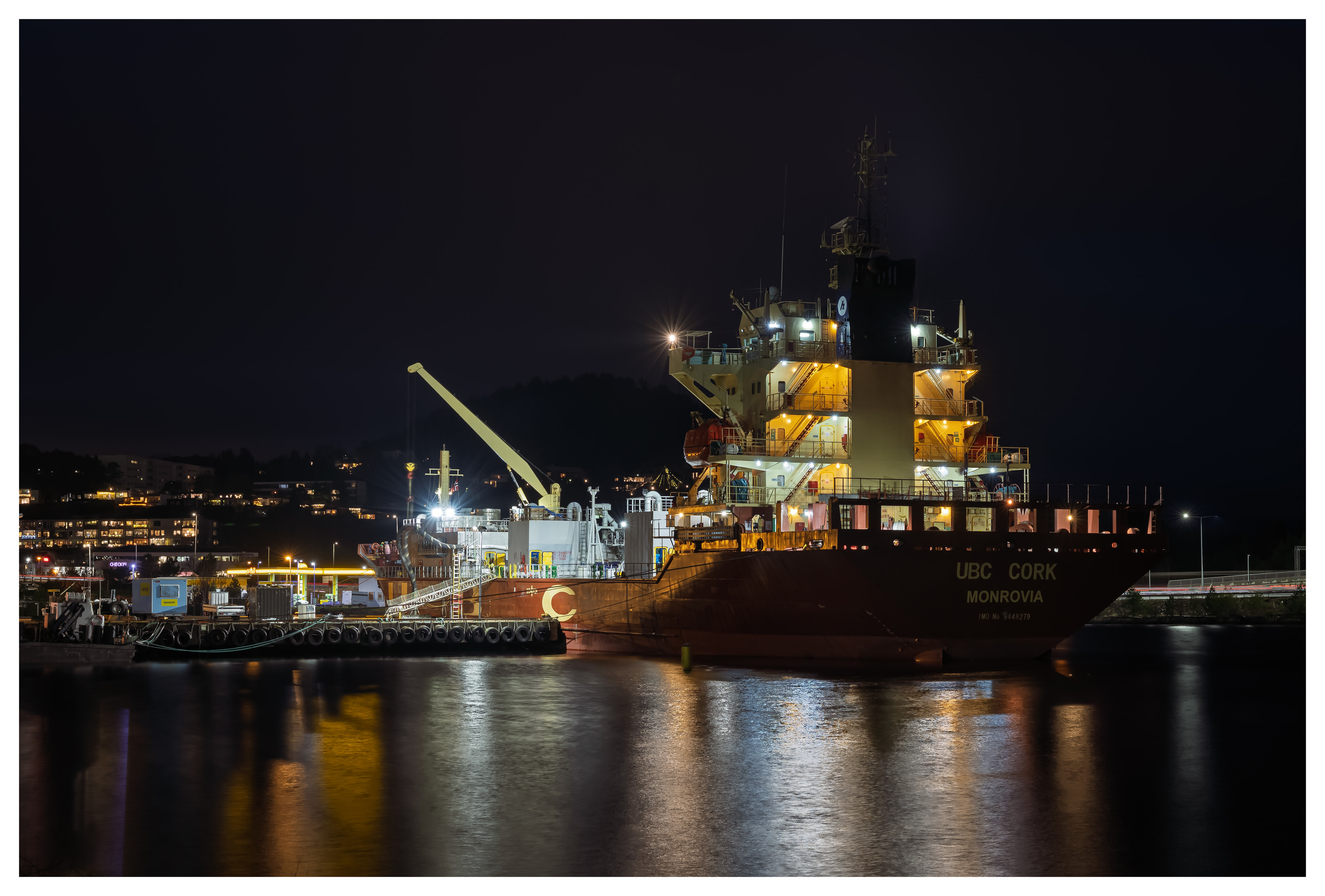 Cargo ship lit up at night in harbor, with bright deck lights reflecting across the dark water.