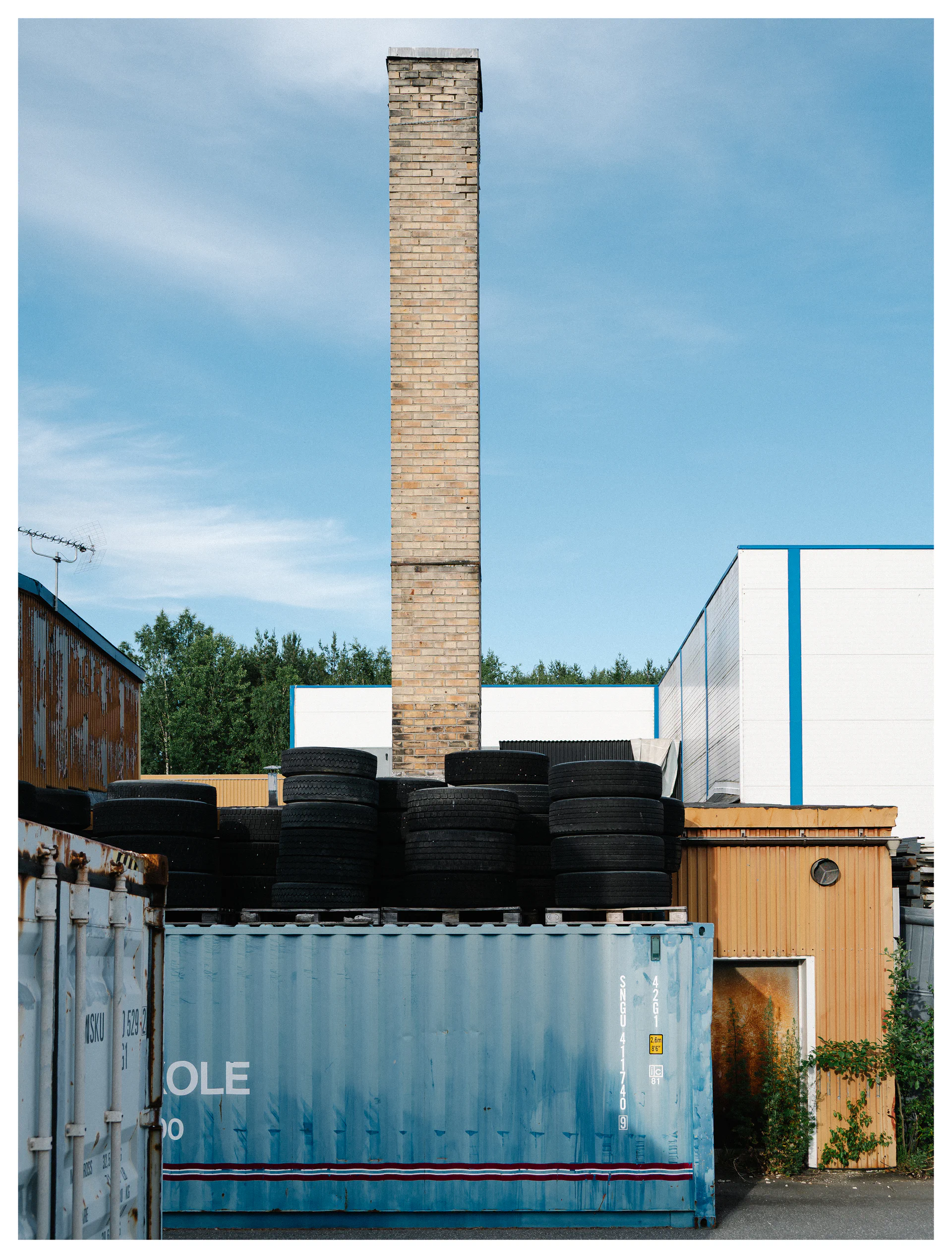 Industrial yard with stacked truck tires, shipping containers, and a tall brick chimney rising against a blue cloudy sky.