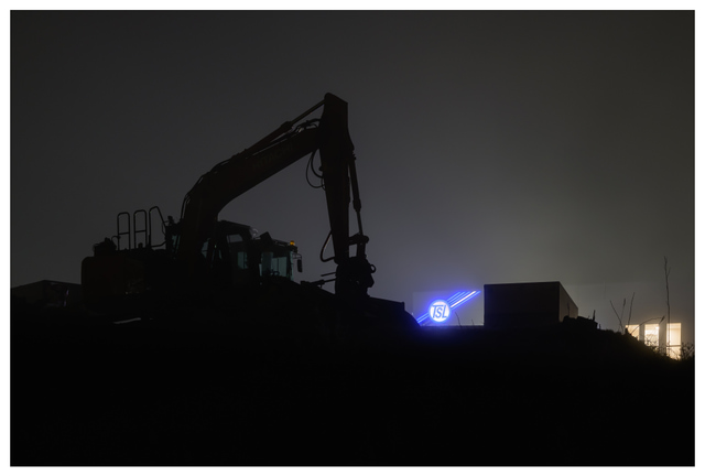Silhouette of an excavator at a nighttime construction site with industrial structures and soft illuminated background lighting.