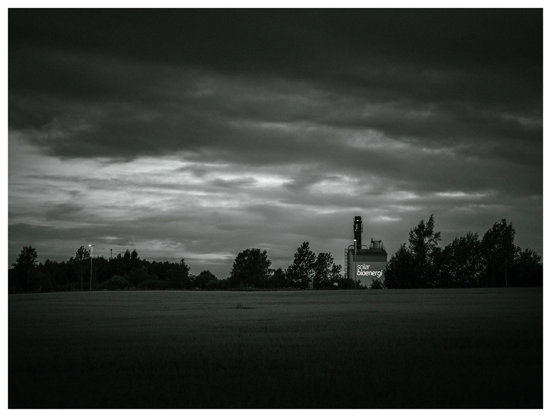 Black and white photograph of an industrial bioenergy plant beyond a field beneath a dramatic overcast sky.