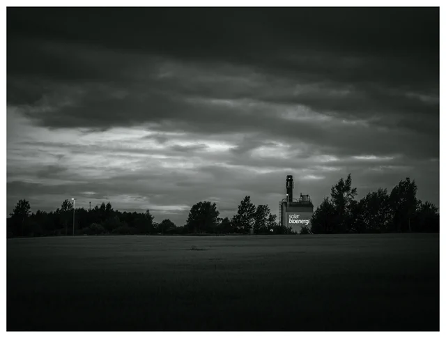 Black and white photograph of an industrial bioenergy plant beyond a field beneath a dramatic overcast sky.