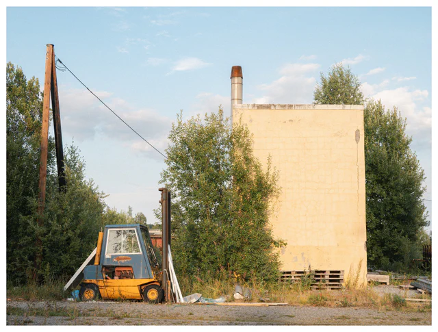 Weathered yellow forklift parked beside an overgrown industrial building in warm evening light.