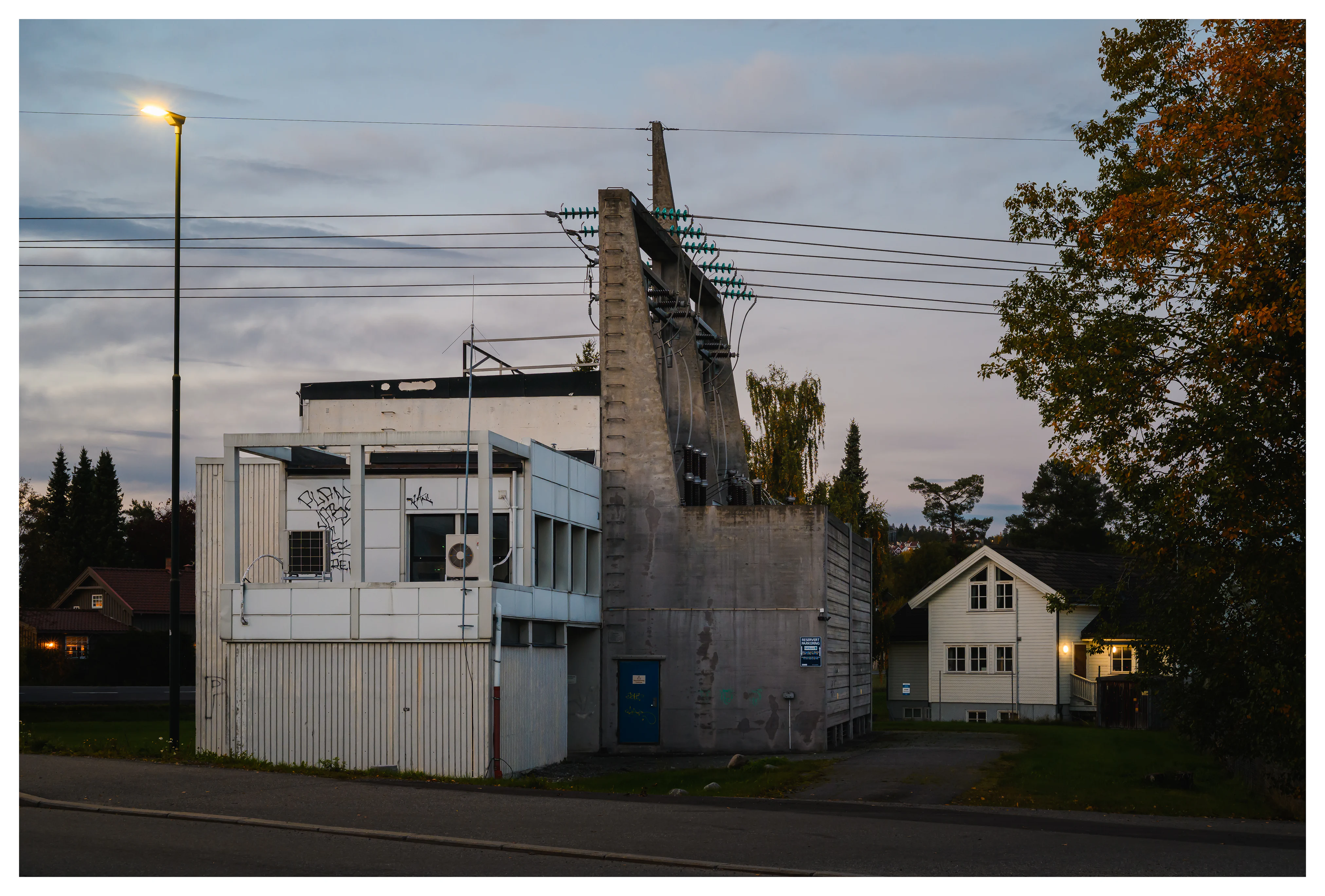 A small electrical substation photographed at dusk, surrounded by houses and trees, its cables and concrete structures faintly lit by evening light.