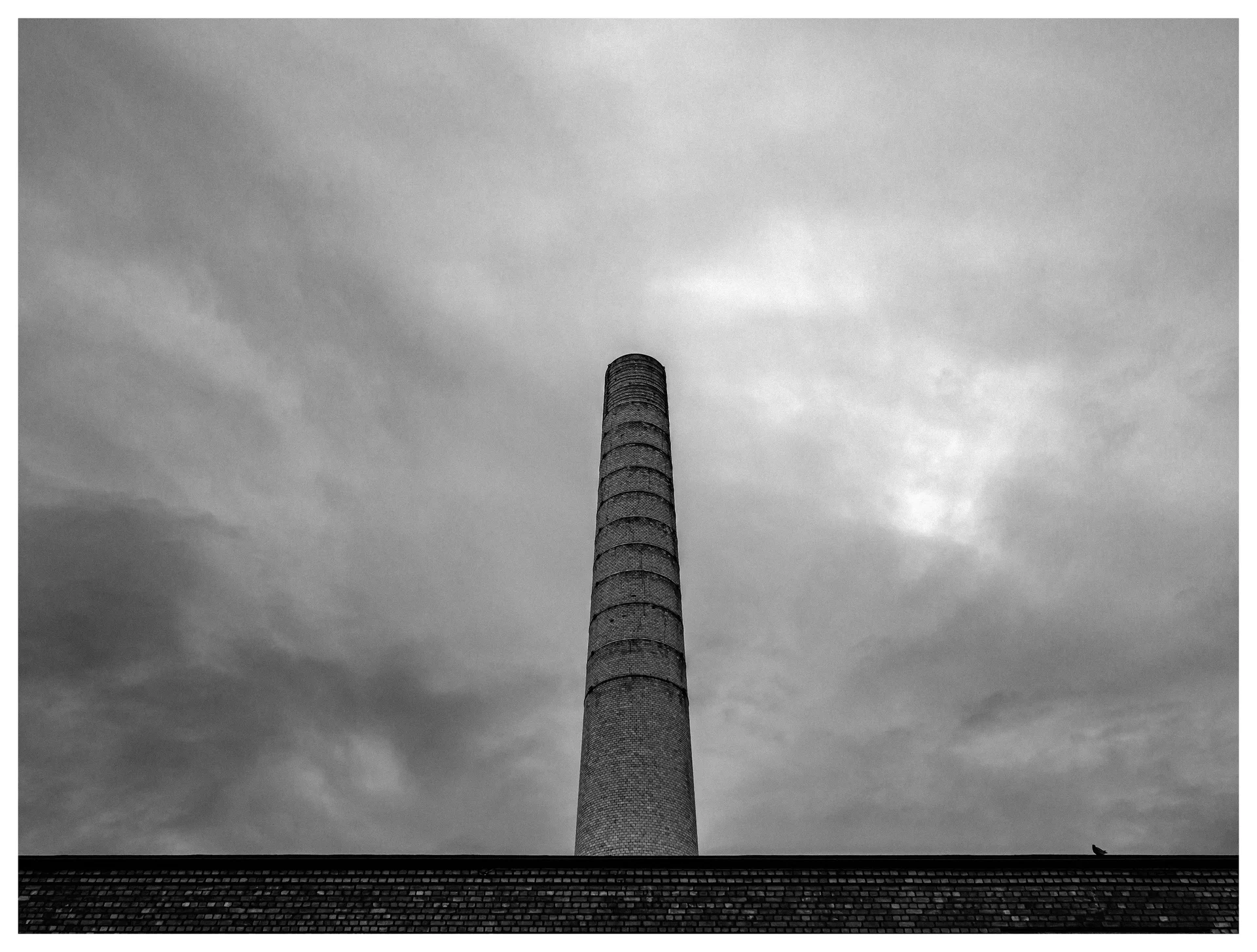 Tall brick industrial chimney rising above a factory roof under dramatic clouds, black and white photograph.