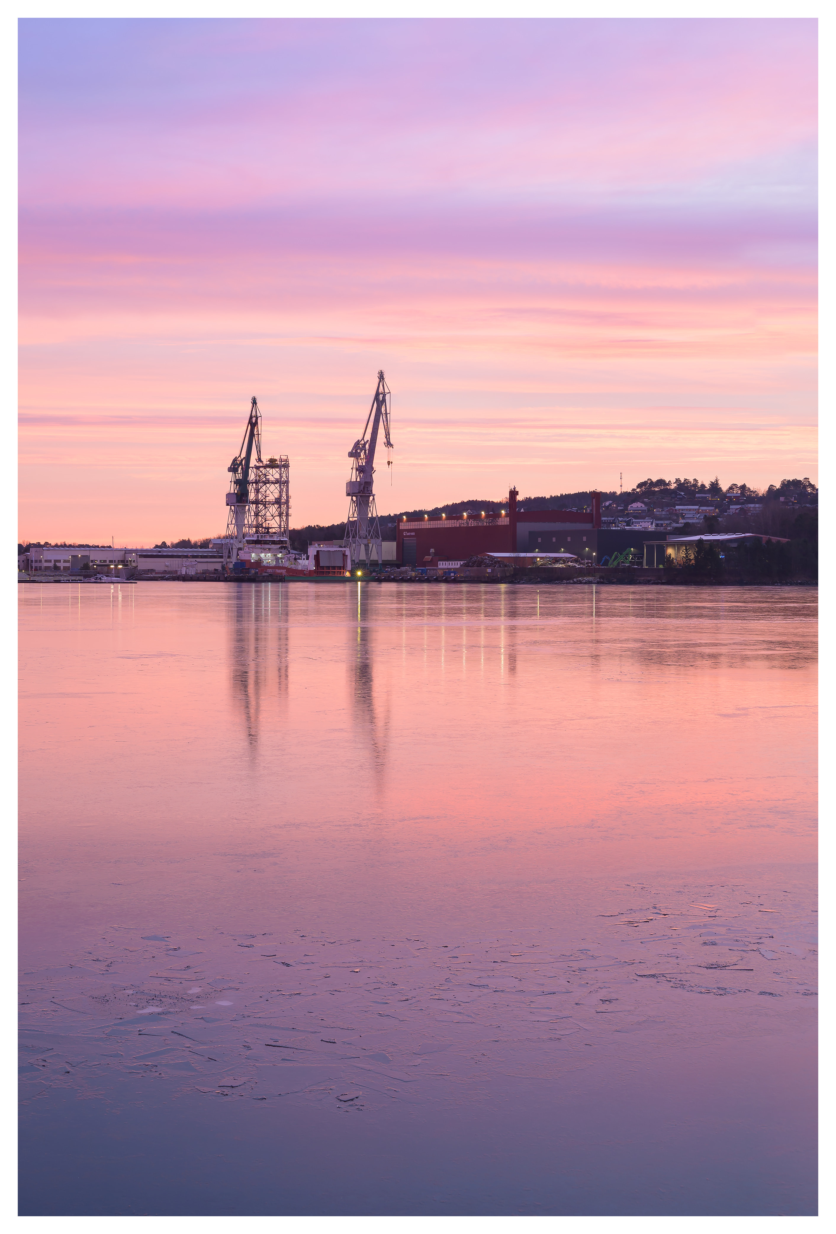 Industrial shipyard cranes reflected in frozen water under a soft pastel evening sky.