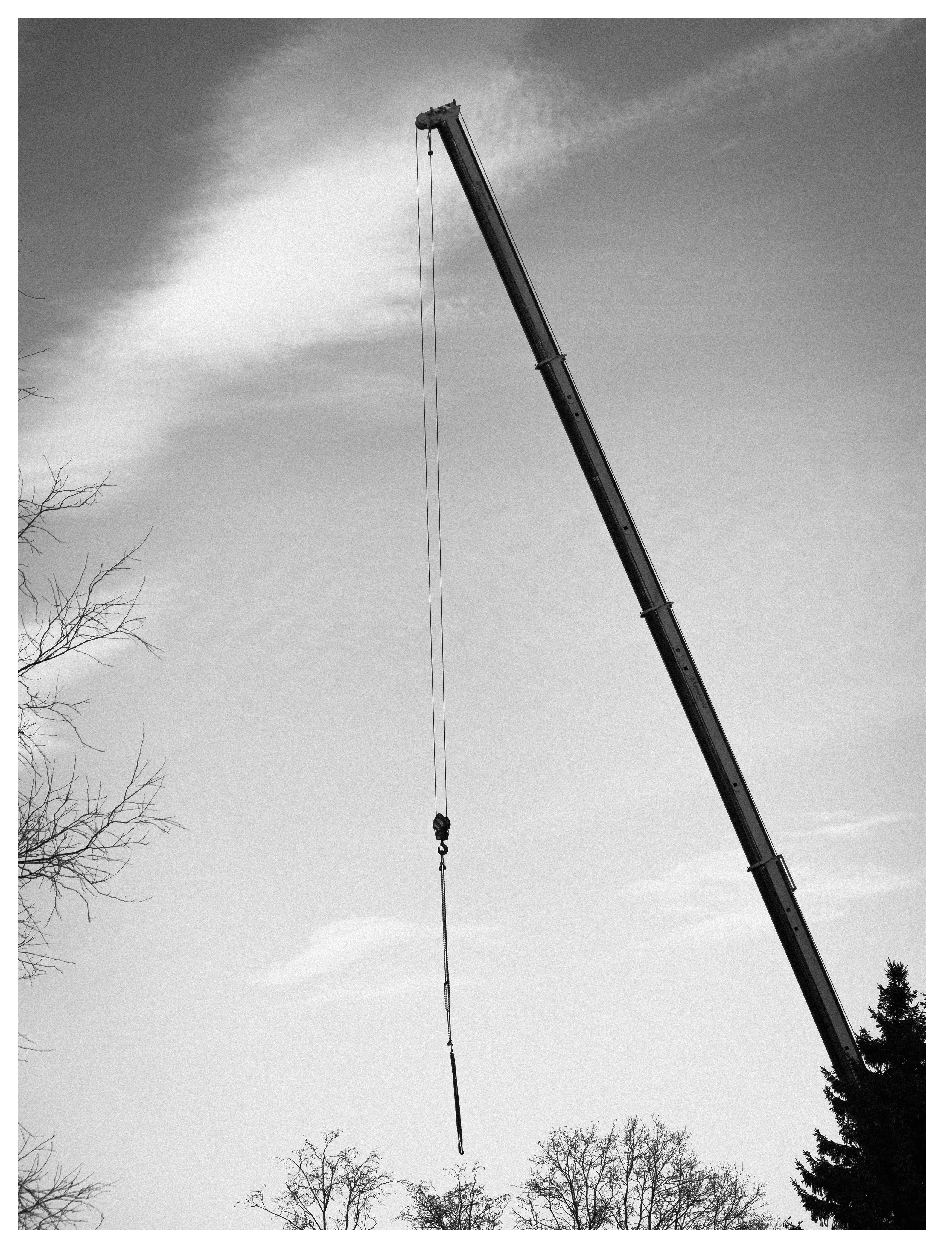 Black and white low-angle photograph of a mobile crane boom extending diagonally across a cloudy sky, with a hanging hook and cable visible, surrounded by bare winter trees.