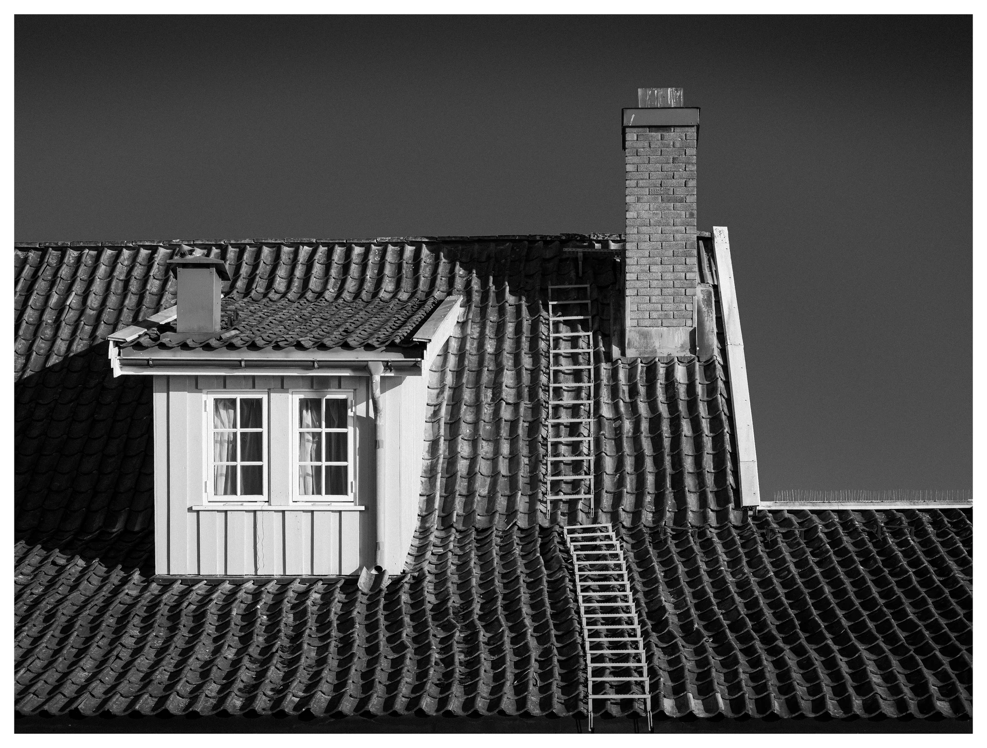 Black and white photograph of a tiled rooftop with a dormer window, chimney and ladder leading across the roof beneath a clear sky.