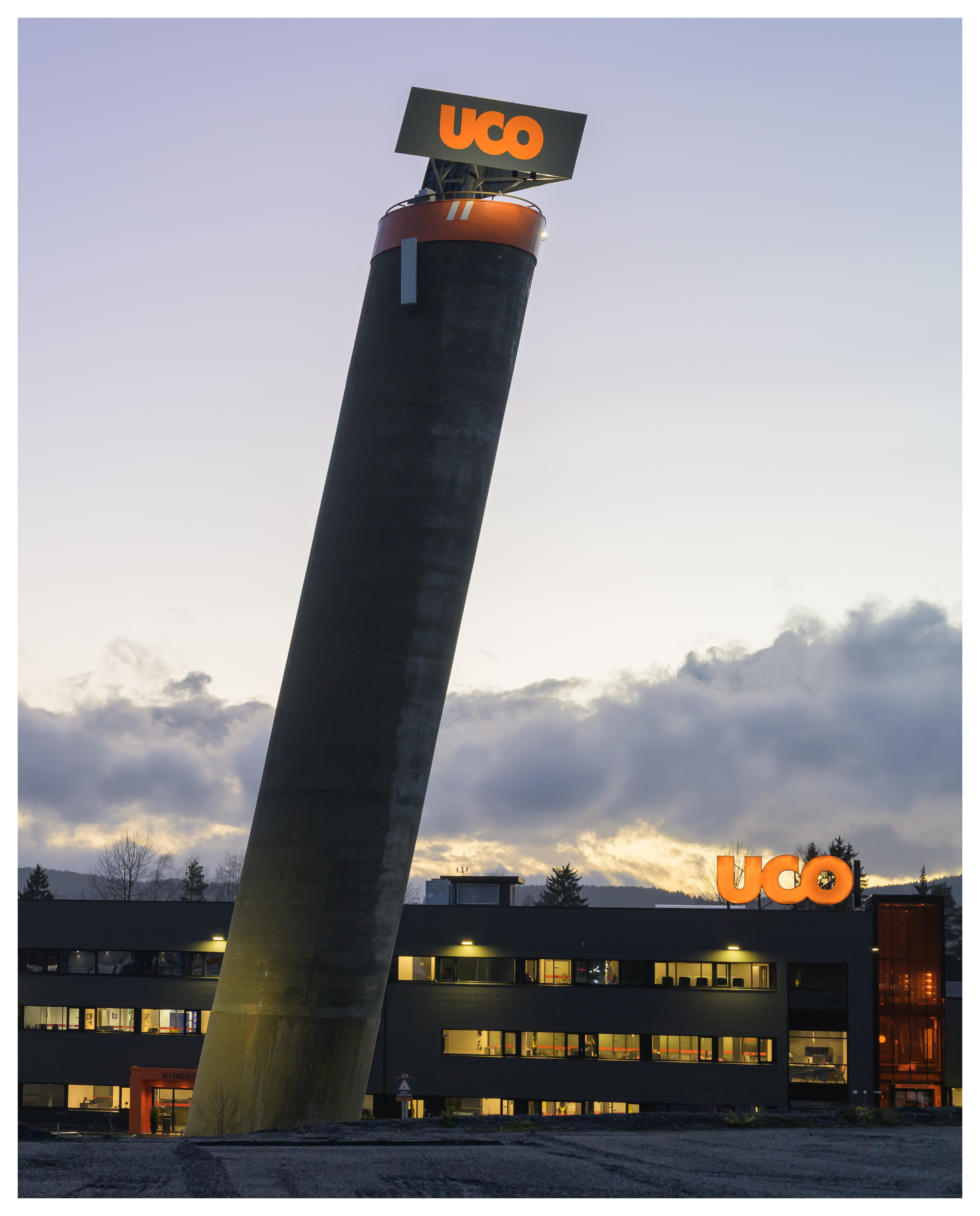 A tilted concrete tower marked with the glowing orange UCO logo rises beside a modern industrial building at dusk, under a pale evening sky.