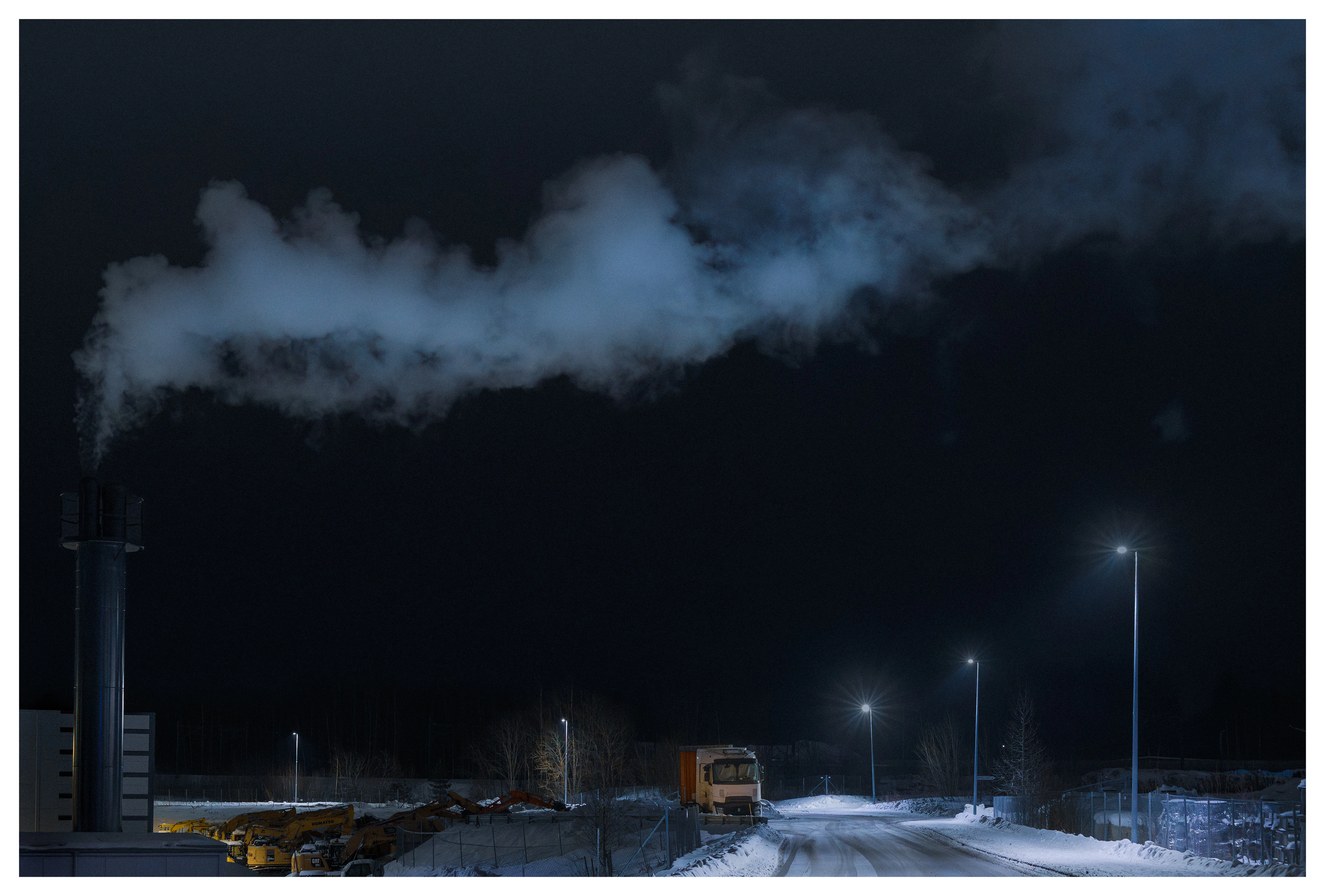 Snowy industrial road at night illuminated by street lights, with steam drifting across the sky from a chimney and a parked truck in a quiet construction area.