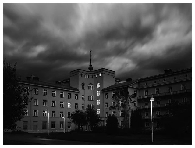Black and white photograph of a large historic building beneath dramatic storm clouds, with a central tower and illuminated windows.