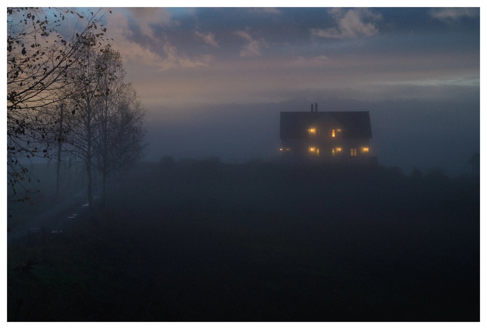 A misty rural evening with a solitary house glowing warmly through the fog, evoking a feeling of homecoming and longing.