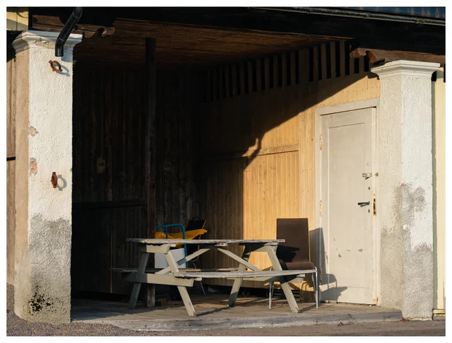 Old wooden porch with a picnic table, mismatched chairs and a slightly open door in warm afternoon sunlight.