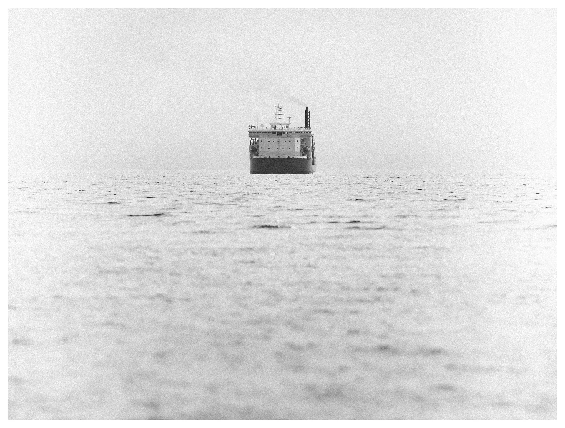 Black and white photograph of a cargo ship on the distant horizon, surrounded by calm open water beneath a bright, hazy sky.
