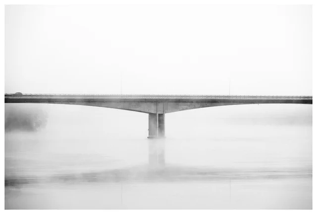 Minimalist black-and-white photograph of a bridge disappearing into morning fog, with a partially visible car crossing at the far left.