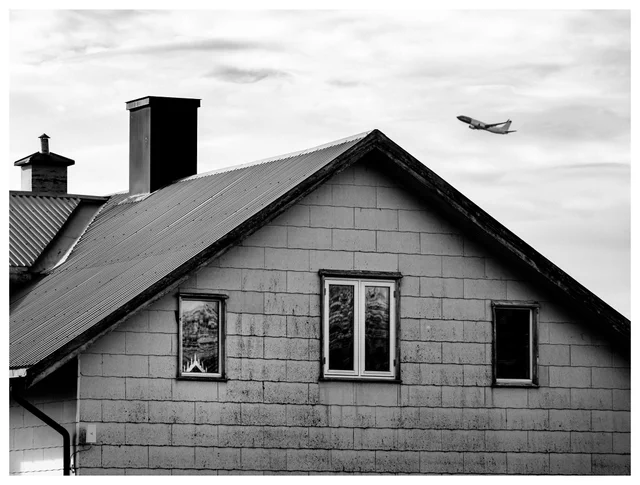 Black-and-white photograph of a worn house with a lit seven-light candle decoration in the window and an airplane passing overhead in the cloudy sky.
