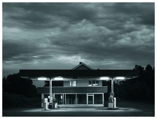 Empty gas station illuminated at night beneath a dramatic cloudy sky, with fuel pumps and a dark building surrounded by trees.