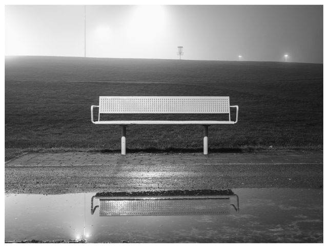 Black and white photo of an empty bench reflected in a puddle on a foggy night, with soft lights and a distant disc golf basket.