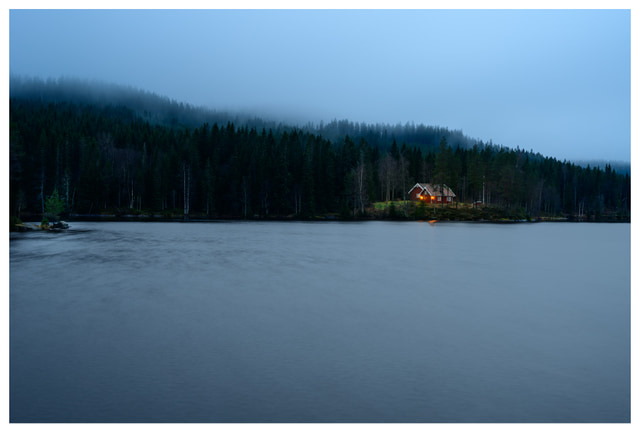 Foggy evening at Ørfiske lake with a softly lit cabin across calm water, surrounded by forested hills in Nordmarka, Norway