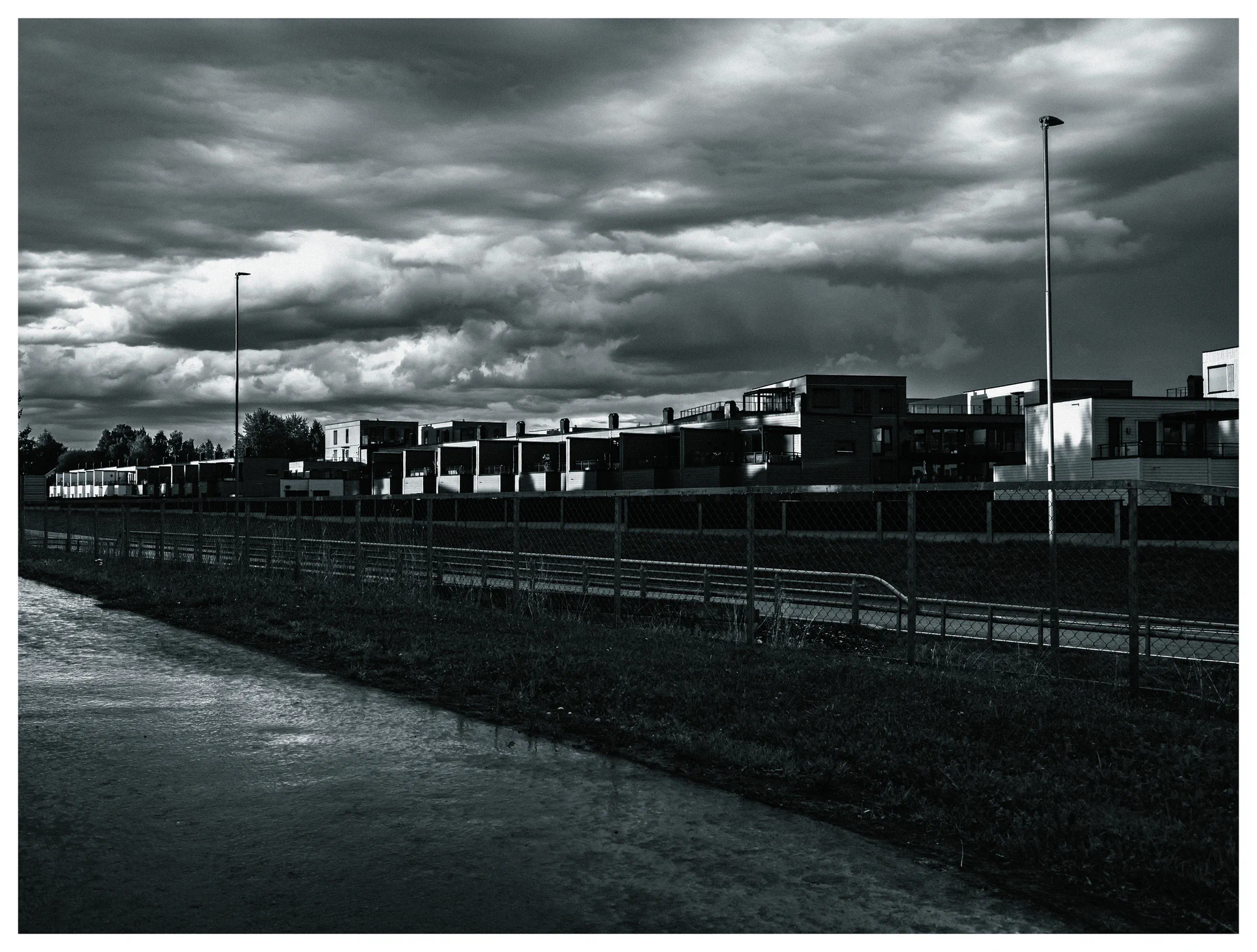 Black and white photograph of modern buildings and streetlights beneath dramatic storm clouds, with wet pavement and fencing in the foreground.