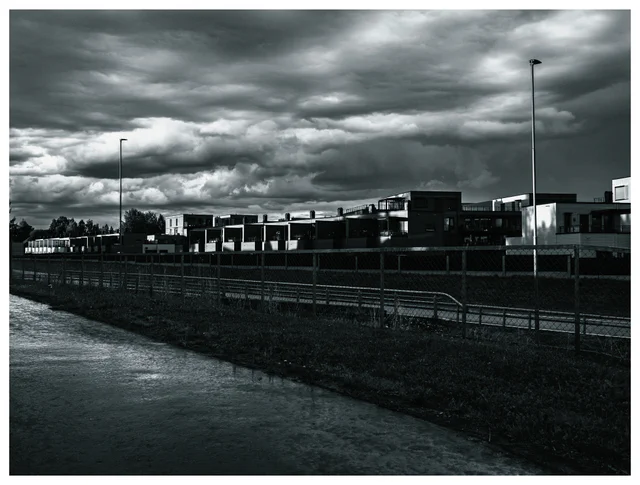 Black and white photograph of modern buildings and streetlights beneath dramatic storm clouds, with wet pavement and fencing in the foreground.