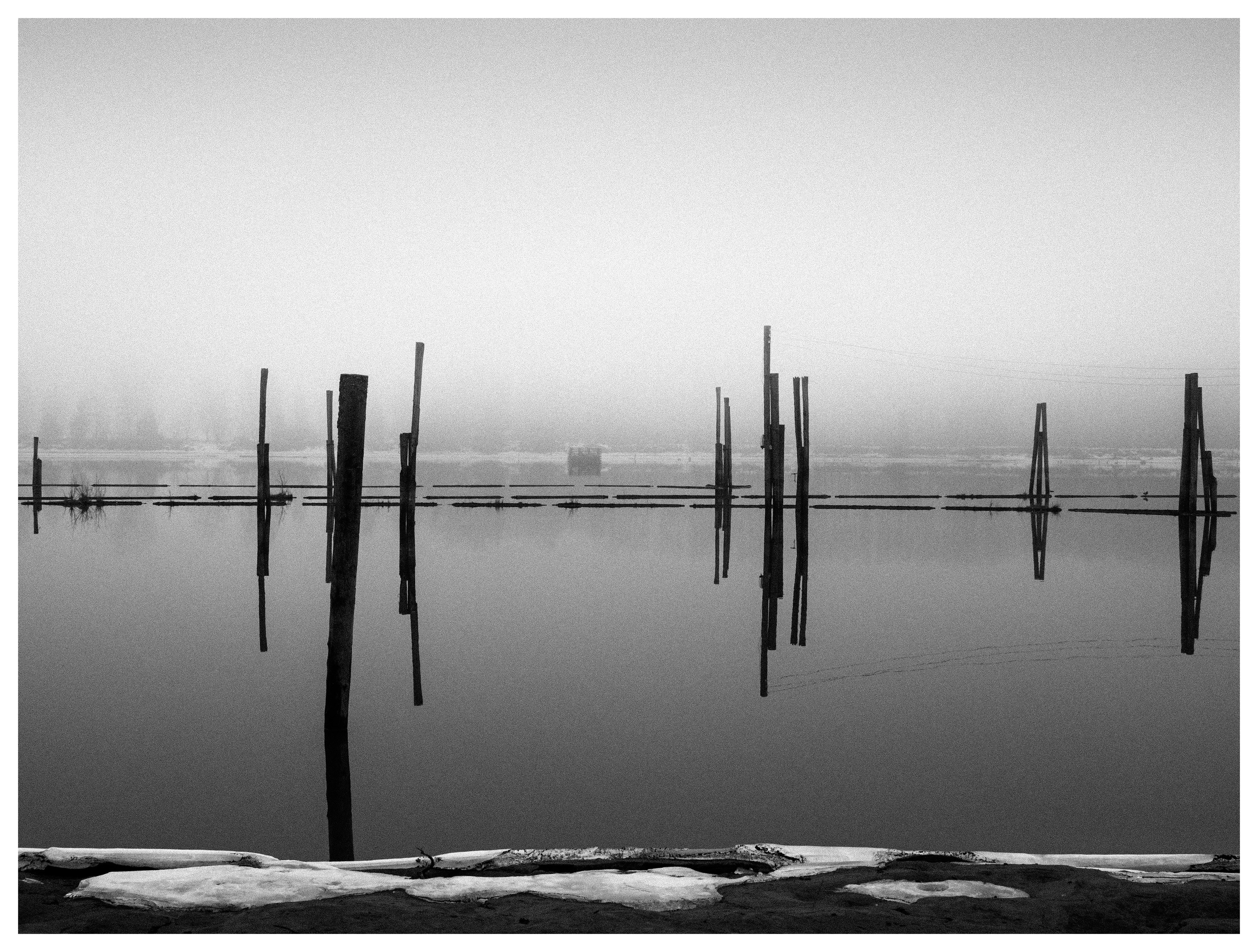 Black and white minimalist fine art photography of historic timber booms and wooden pilings at Fetsund Lenser, reflecting in the calm, foggy waters of the Glomma river in Norway.