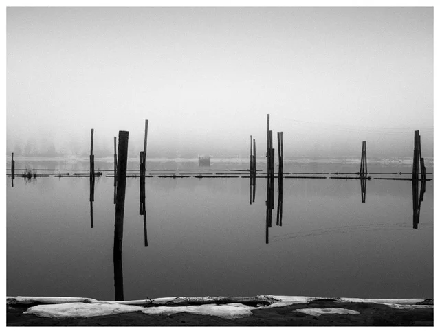 Black and white minimalist fine art photography of historic timber booms and wooden pilings at Fetsund Lenser, reflecting in the calm, foggy waters of the Glomma river in Norway.