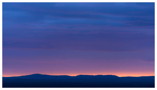Minimalist landscape at dusk with a dark distant ridge beneath a vast blue and violet sky, with a narrow band of warm light along the horizon.