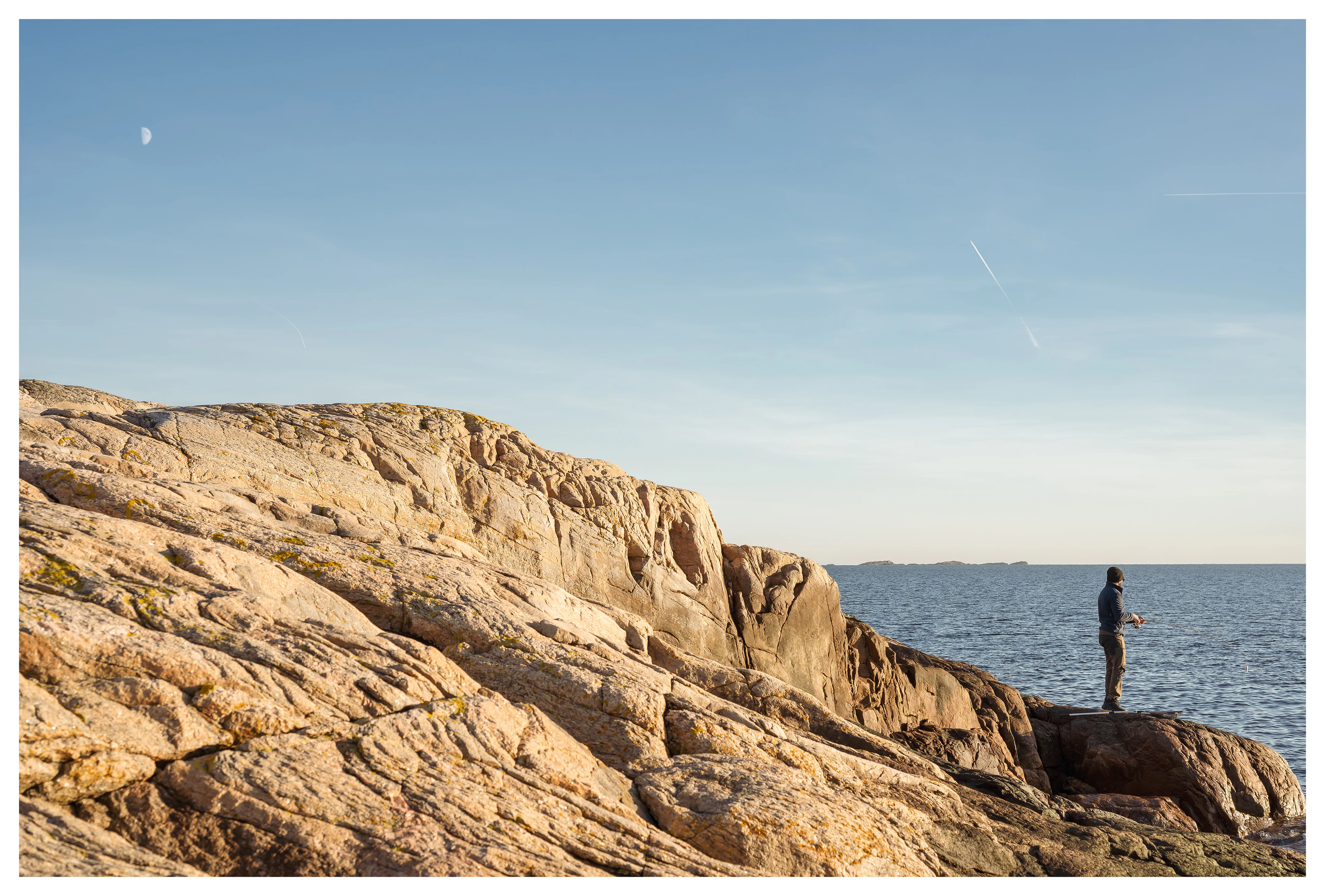 Coastal landscape at sunset with rugged rocks and a lone person by the sea, capturing calm Scandinavian shoreline light and a quiet sense of space.