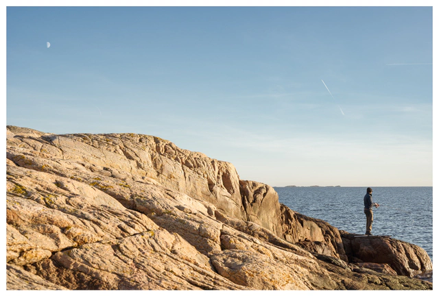 Coastal landscape at sunset with rugged rocks and a lone person by the sea, capturing calm Scandinavian shoreline light and a quiet sense of space.