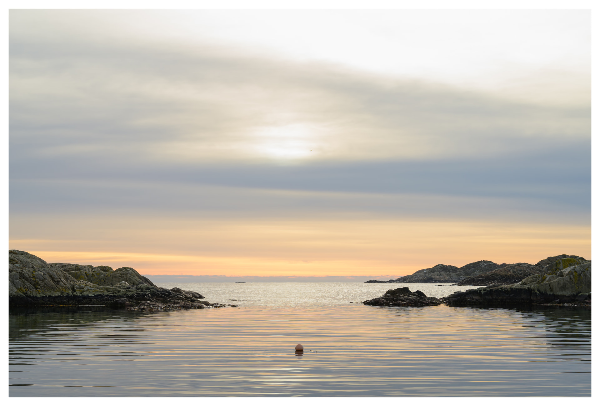 Calm bay at sunset with soft layered clouds, rocky coastline, gentle ripples, and a single buoy floating in the water.