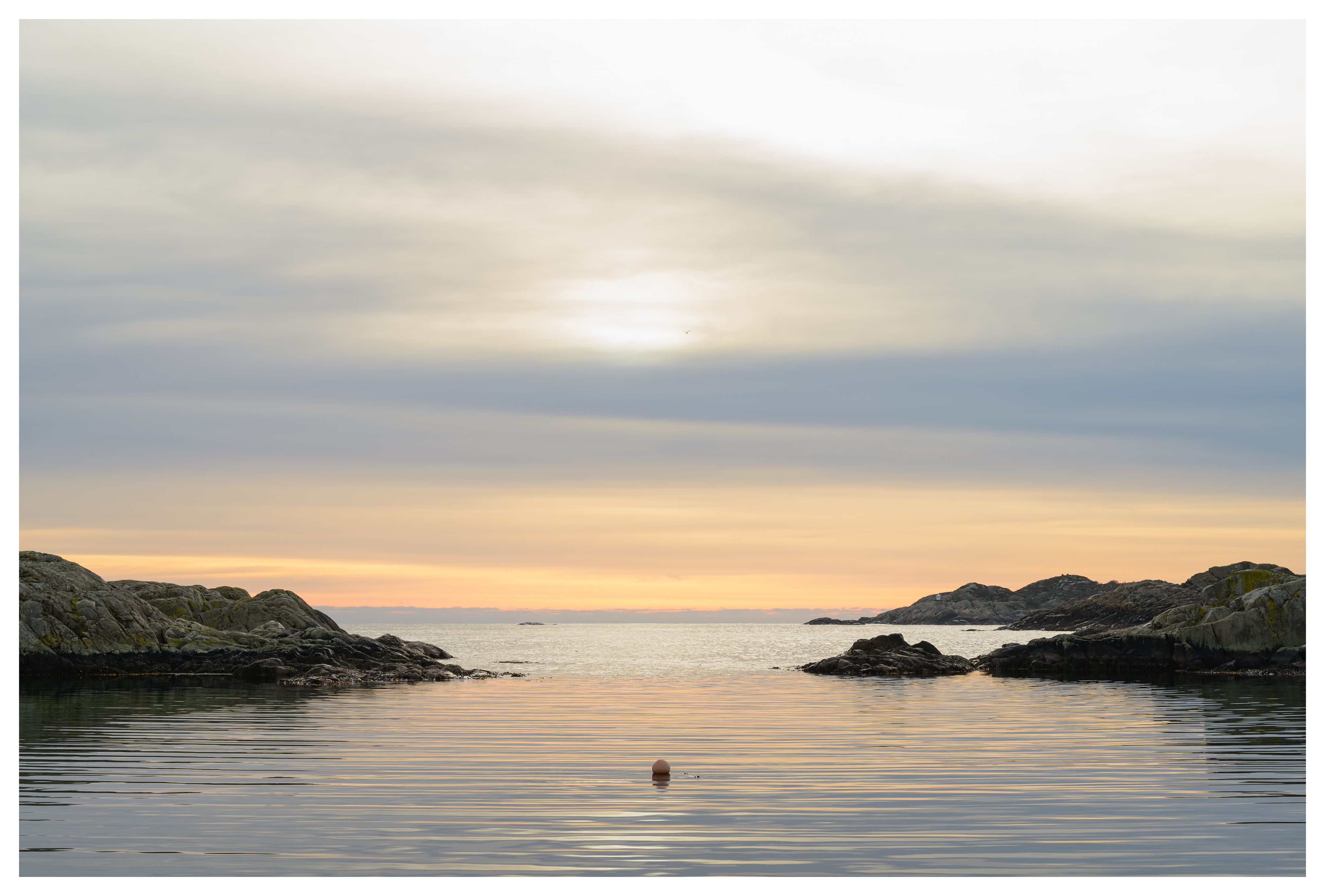 Calm bay at sunset with soft layered clouds, rocky coastline, gentle ripples, and a single buoy floating in the water.