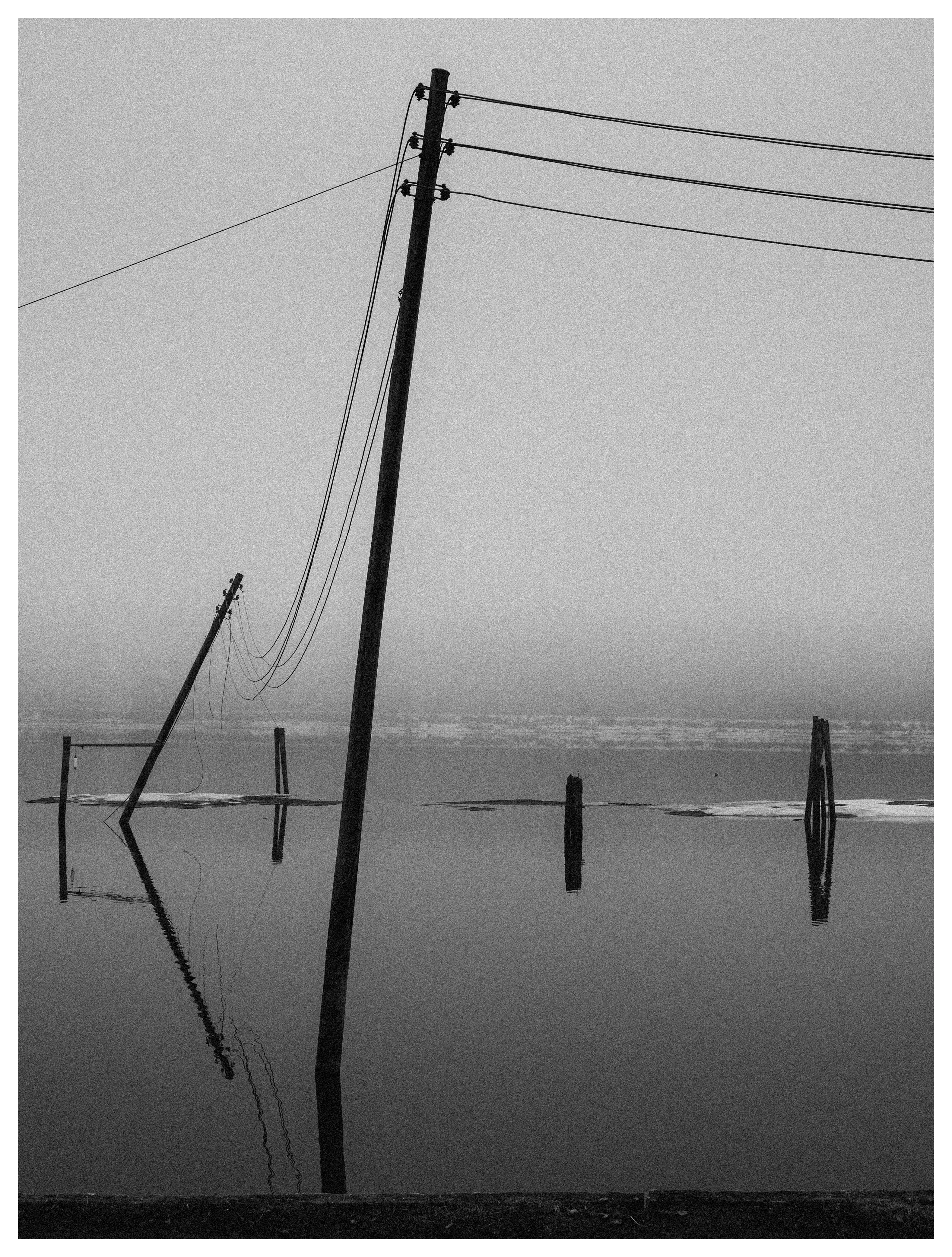 Black and white photo of leaning power poles and power lines reflected in still floodwater under a foggy sky.