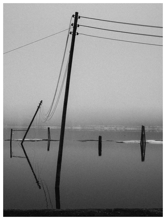 Black and white photo of leaning power poles and power lines reflected in still floodwater under a foggy sky.