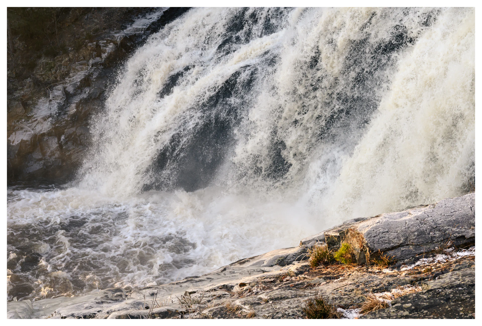 Powerful waterfall crashing over dark rock, with swirling white water and sunlit stone in the foreground.