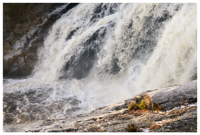 Powerful waterfall crashing over dark rock, with swirling white water and sunlit stone in the foreground.
