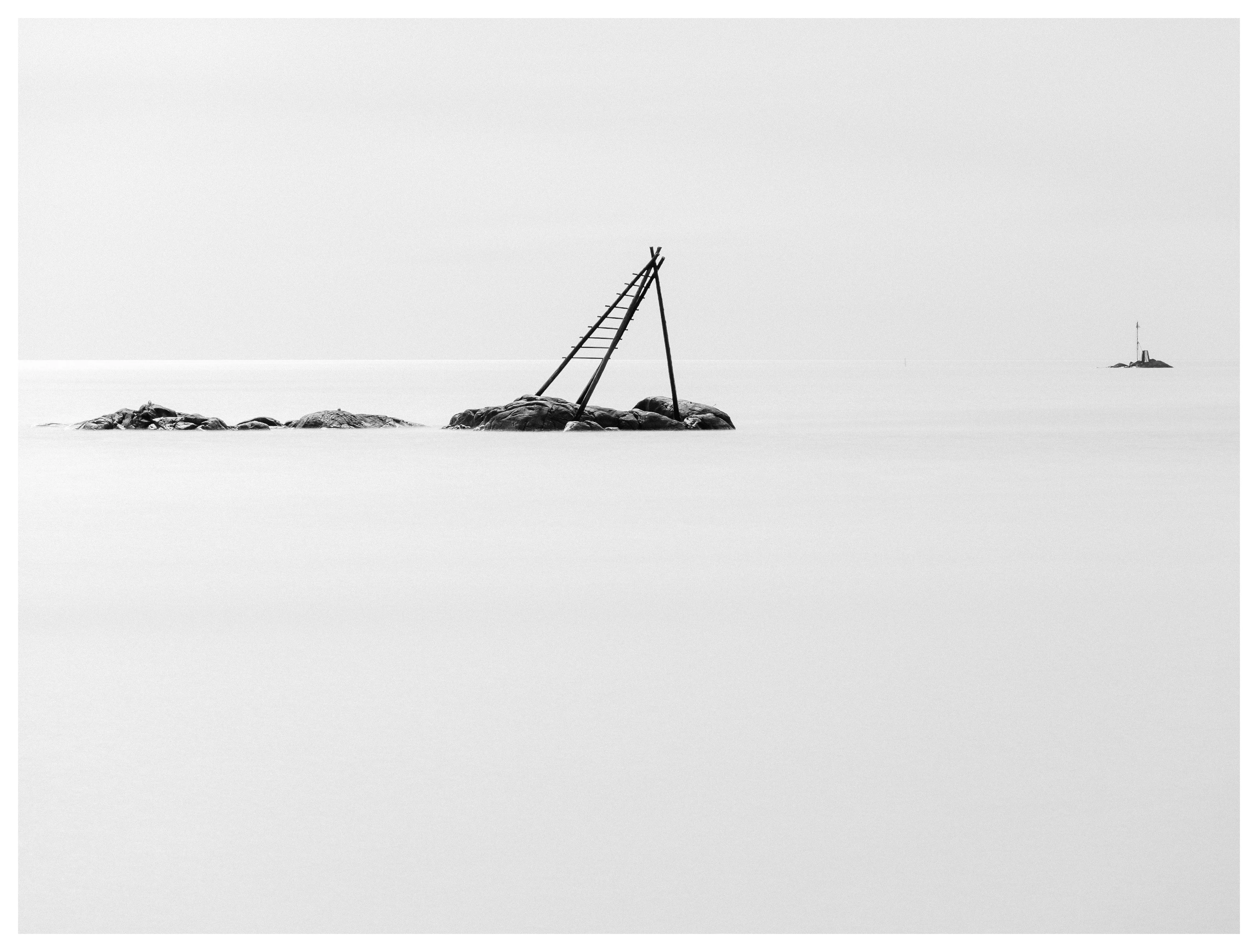 Black and white minimalist seascape with tilted wooden structure on rocky islet in calm water.