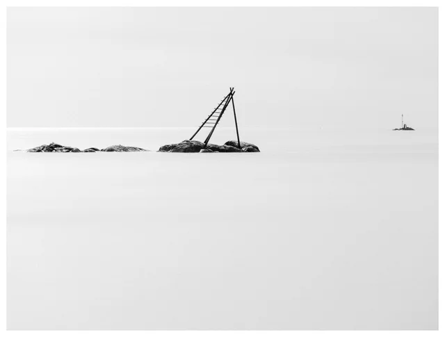 Black and white minimalist seascape with tilted wooden structure on rocky islet in calm water.